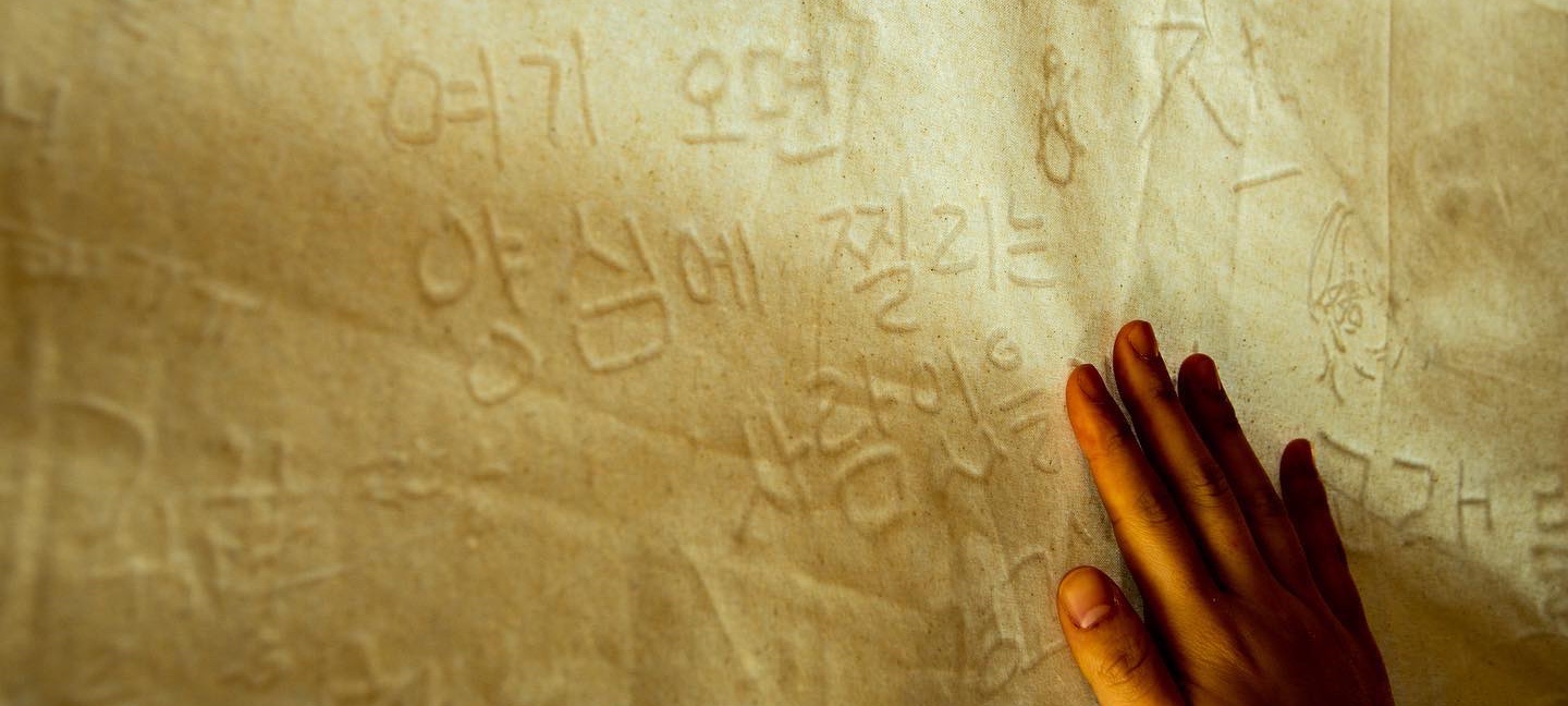 전시장에 걸린 천에 사람들이 손으로 쓴 글씨가 가득하고, 그 위에 손 하나가 가만히 놓여 있다.